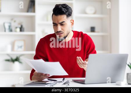 Ein besorgter arabischer Mann, der mit der Hand auf Dokumente blickt, die zu Hause mit Laptop am Tisch sitzen Stockfoto