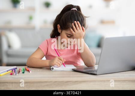 Mädchen aus dem Mittleren Osten im Pferdeschwanz, die Stirn auf der Hand gelehnt und auf Notizbuch geschrieben hat, während sie zu Hause am Schreibtisch im Wohnzimmer sitzt Stockfoto