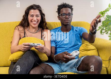 Enttäuschter Schwarzer Mann, der die Hand nach vorne zeigt, während er ein Videospiel mit lachender kaukasischer Freundin spielt, die zu Hause auf der Couch sitzt Stockfoto