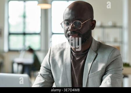 Lächelnder schwarzer Glatzkopf mit Brille, der am Laptop arbeitet und am Schreibtisch im Büro sitzt Stockfoto