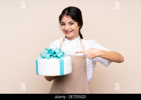 Hispanische Konditorin zeigt auf Kuchen, der vor beigefarbenem Hintergrund im Studio steht Stockfoto