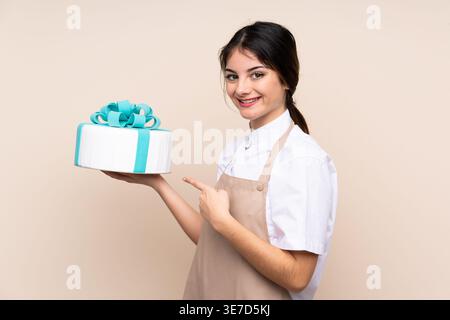 Hispanische Konditorin zeigt auf Kuchen, der seitlich vor beigem Hintergrund im Studio steht Stockfoto