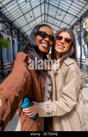Schwarze und kaukasische Freundinnen tragen Trenchcoat und Sonnenbrille und machen Selfies, während sie tagsüber in der Stadt stehen Stockfoto