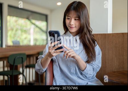 Thailändische Frau, die ihr Smartphone benutzt, während sie am Holztisch im Café sitzt Stockfoto