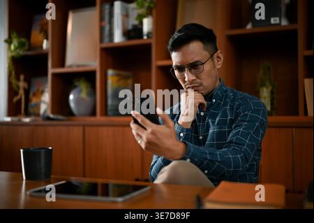 Seriöser asiatischer Geschäftsmann mit blauen karierten langen Ärmeln und smLifestylephone, während er im Büro mit der Hand am Kinn sitzt Stockfoto