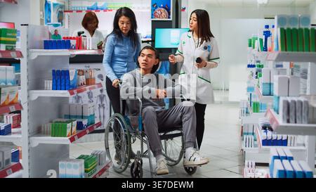 Junger Mann Rollstuhlfahrer spricht mit Apotheker über Medikamente, fragt nach verschreibungspflichtigen Behandlungen oder Medikamenten. Behinderte und Betreuer, die Nahrungsergänzungsmittel und Vitamine in der Apotheke erhalten. Stockfoto