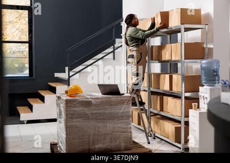 Lagermitarbeiter, der sich ein Metallregal ansieht und während des Lagerbestands den Karton herausnimmt. afroamerikaner, der Kundenaufträge vorbereitet, an Paketen im Lager arbeitet Stockfoto