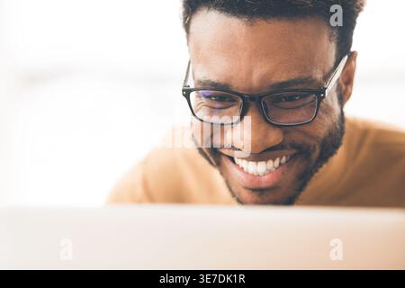 Ein lächelnder schwarzer Mann mit Brille, der auf den Laptop im Büro am Tisch sitzt Stockfoto