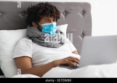 Kranker asiatischer Mann mit lockigem Haar, der Schal um den Hals und Gesichtsmaske mit Laptop trägt, während er zu Hause im Bett lag Stockfoto