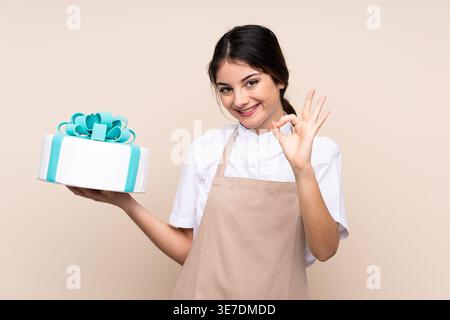 Hispanische Konditorin mit OK-Hand-Schild mit Kuchen, der vor beigem Hintergrund im Studio steht Stockfoto
