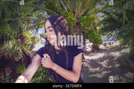 Hispanische Touristin, die Insektenschutzmittel am Arm aufträgt und am sonnigen Sommertag neben Palmen steht Stockfoto