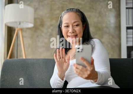 Lächelnde ältere asiatische Frau, die die Hand winkt und auf dem Smartphone Video auf dem Sofa sitzt Stockfoto