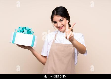 Hispanische Konditorin zeigt ein Friedensschild mit Kuchen, der vor beigefarbenem Hintergrund im Studio steht Stockfoto