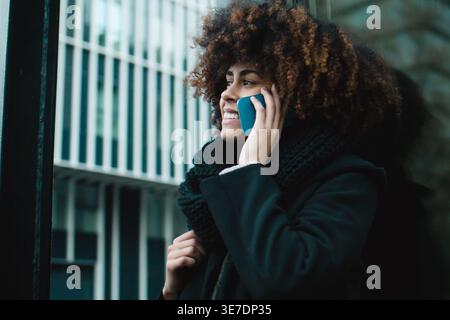 Lächelnde Mixed Race-Frau mit Afro, die eine schwarze warme Jacke und einen Schal trägt und einen Anruf macht, während sie im Winter auf der Straße steht Stockfoto