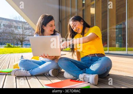 Fröhliche asiatische und kaukasische Studentin, die zusammen mit einem Laptop sitzt und mit überkreuzten Beinen auf einer Holzterrasse vor dem Universitätsgebäude auf der sonnigen... Stockfoto
