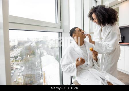 Lächelnde schwarze Frau in weißem Gewand, die die Lippen des Schwarzen Freundes berührt, der frühstückt, während er auf dem Stuhl neben dem Fenster im Hotelzimmer sitzt Stockfoto