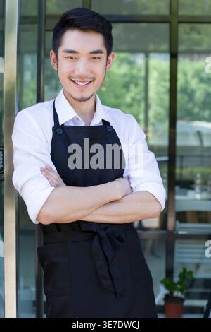 Ein chinesischer Mann mit Schürze, der sich an der Tür mit gekreuzten Armen im Coffee Shop beugt Stockfoto