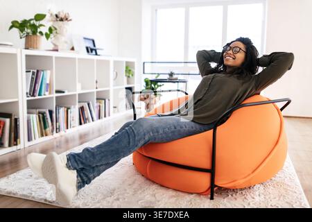 Schwarze Frau mit lockigen Haaren, die Brille trägt, sitzt mit Händen hinter dem Kopf und geschlossenen Augen auf Sitzsack im Wohnzimmer zu Hause Stockfoto