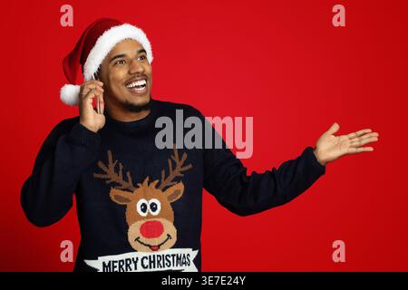 Fröhlicher Schwarzer Kerl trägt Weihnachtsmütze und Pullover mit Hirsch, der wegschaut, während er mit offener Hand vor rotem Hintergrund steht... Stockfoto