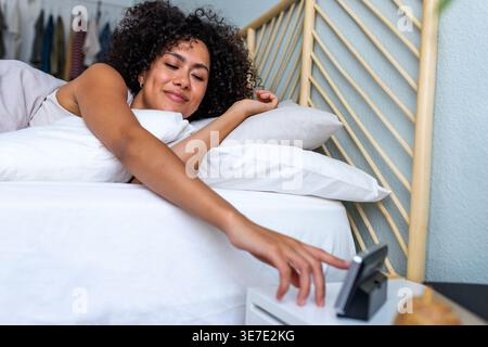 Lächelnde hispanische Frau, die den Wecker zum Ausschalten erreicht, während sie zu Hause auf dem Bett liegt Stockfoto