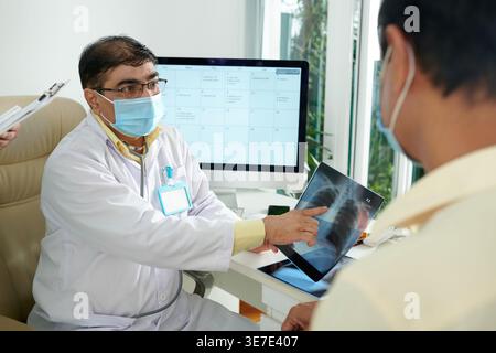 Indischer männlicher Arzt mit Gesichtsmaske, der dem asiatischen männlichen Patienten Röntgenergebnisse zeigt, während er am Tisch in der Klinik sitzt Stockfoto