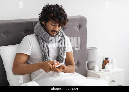 Kranker asiatischer Mann mit lockigen Haaren, der einen Schal um den Hals trägt und Pillen nimmt, während er zu Hause im Bett sitzt Stockfoto