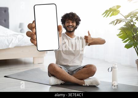 Asiatischer Mann mit lockigen Haaren, der Smartphone-Bildschirm zeigt, während er mit überkreuzten Beinen auf Yogamatte im Wohnzimmer zu Hause sitzt Stockfoto