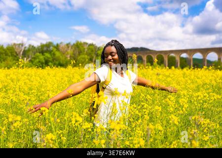 Lächelnde schwarze Frau mit Wendungen, die mit ausgestreckten Armen auf einem Feld aus gelben Blumen steht Stockfoto