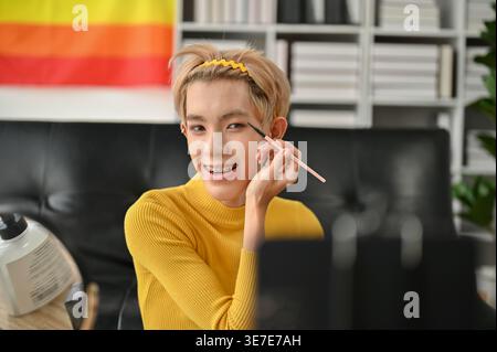 Lächelnder asiatischer schwuler Mann, der Lidschatten während der Aufnahme aufträgt Make-up Tutorial sitzend auf dem Boden in der Nähe des Sofas zu Hause Stockfoto