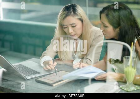 Seriöse asiatische Geschäftsfrau mit Eingabestift, die ein Tablett zum Blättern hält, diskutiert Details mit kaukasischen Kollegen, die tagsüber zusammen sitzen Stockfoto