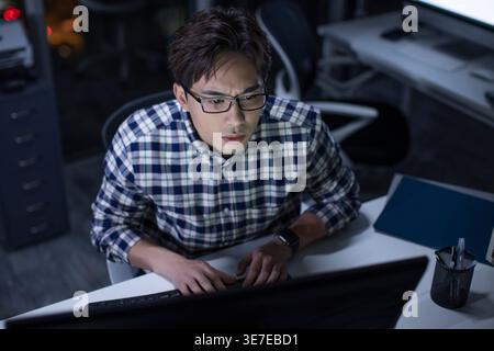 Seriöser chinesischer Mann mit Brille, der karierte lange Ärmel trägt und nachts spät im Büro arbeitet Stockfoto