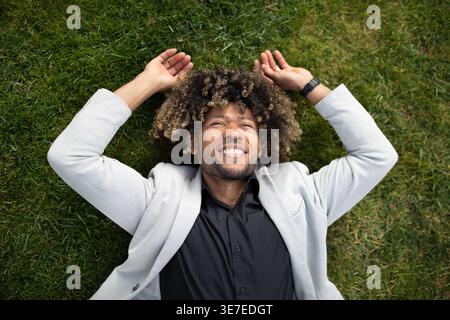 Lächelnder hispanischer Geschäftsmann mit lockigem Afro-Anzug, der tagsüber auf grünem Rasen im Park liegt Stockfoto