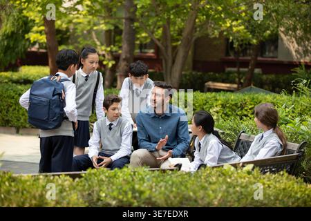 Verschiedene Gruppe von Schülern mit kaukasischen männlichen Lehrern, die tagsüber auf der Bank im Schulpark sitzen Stockfoto