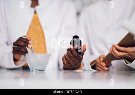 Zugeschnittenes Bild von schwarzen Apothekerinnen, die Pissel und Glasflasche mit medizinischem Inhaltsstoff halten und neben einer japanischen Kollegin stehen... Stockfoto