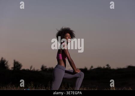 Lächelnde schwarze Athletin mit Afro tragenden Ohrhörern, die wegschaut und beim Stretching mit einem Knie auf dem Feld bei Sonnenaufgang kniend macht Stockfoto
