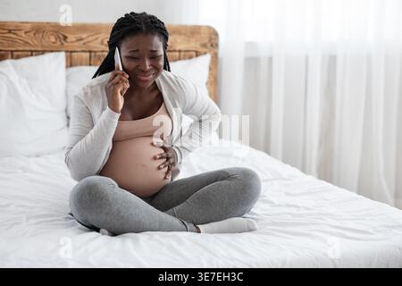 Schwarze schwangere Frau, die einen Anruf hat, der den Bauch berührt und unter Schmerzen leidet, während sie im Schlafzimmer zu Hause auf dem Bett sitzt Stockfoto