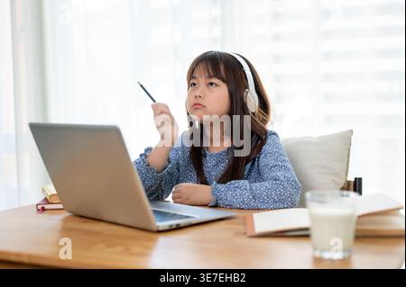 Asiatisches Mädchen mit Kopfhörern, die einen Stift halten, und Online-Kurs zu Hause am Tisch sitzen Stockfoto