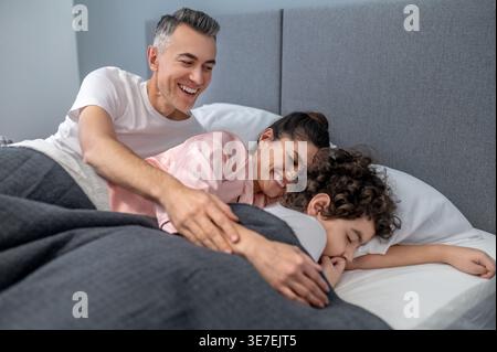 Arabischer Junge mit lockigem Haar, der friedlich mit seinen Eltern lacht, während er zu Hause zusammen auf dem Bett liegt Stockfoto