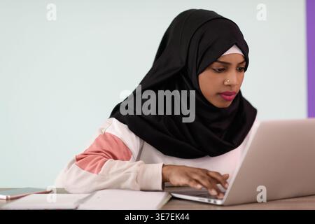 Ernsthafte schwarze muslimische Geschäftsfrau mit Nasenpiercing, die Hijab trägt und am Laptop arbeitet, während sie im Büro am Tisch sitzt Stockfoto