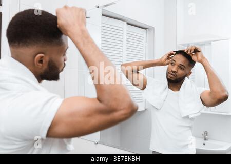 Ernster schwarzer Mann kämmt Haare vor dem Spiegel, der zu Hause im Badezimmer steht Stockfoto