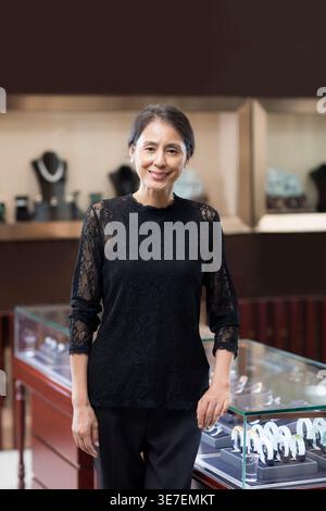 Lächelnde chinesische Käuferin mittleren Alters in schwarzem Kleid, die neben einem Glasvitrine im Juweliergeschäft steht Stockfoto