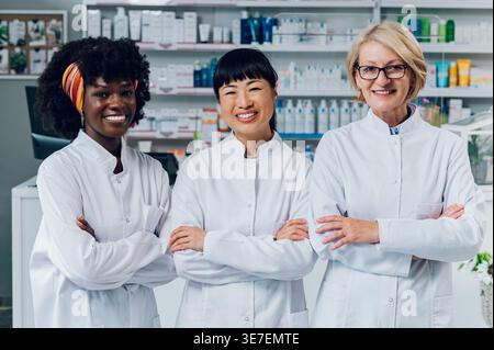 Diverse Gruppe von Apothekerinnen, die mit gekreuzten Armen gegen verschiedene Medizinprodukte in der Apotheke stehen Stockfoto