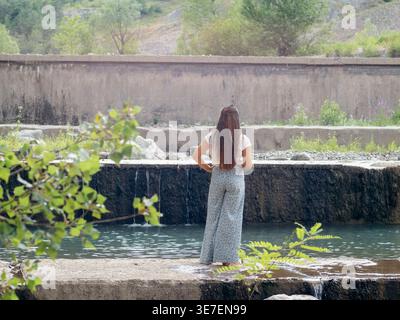 Gemischte Rassenfrau mit Hand auf Taille, die tagsüber auf Beton in der Nähe des Flusses steht Stockfoto