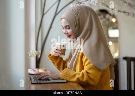 Lächelnde asiatische muslimische Frau, die Hijab trägt und Kaffee trinkt, während sie am Tisch im Café sitzt Stockfoto