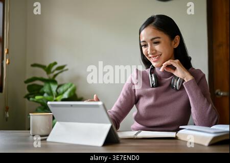 Lächelnde asiatische Frau mit Kopfhörern am Hals, die ein Tablet benutzt, während sie zu Hause mit Büchern am Tisch sitzt Stockfoto