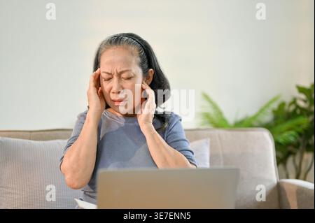 Asiatische Seniorin, die ihre Schläfe berührt und Kopfschmerzen hat, sitzend auf dem Sofa mit Laptop zu Hause Stockfoto