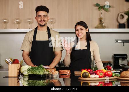 Ein Mann aus dem Nahen Osten und eine hispanische Frau, die ihre Hand winkt, während sie in der Küche steht Stockfoto
