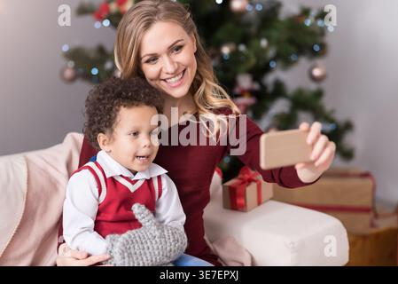 Ein lächelnder Mixed Race Boy macht Selfie mit ihrer Mutter, die zu Hause auf der Couch neben dem Weihnachtsbaum sitzt Stockfoto