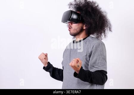 Hispanischer Mann mit lockigem Haar, der im Studio eine VR-Brille mit geballten Fäusten vor weißem Hintergrund trägt Stockfoto