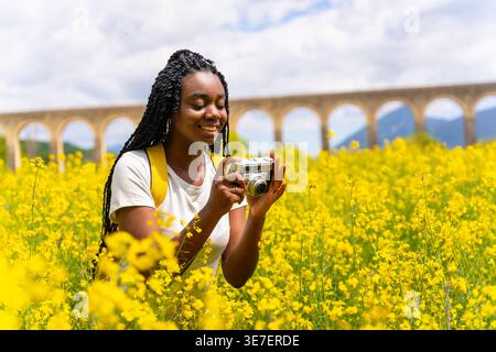 Lächelnde schwarze Frau mit Wendungen, die gelbe Blumen fotografiert, die am sonnigen Tag auf dem Feld stehen Stockfoto